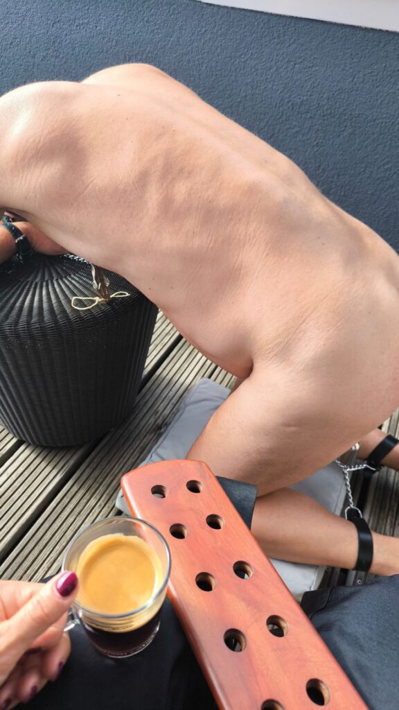 Morning BDSM session in Berlin with submissive kneeling while domina enjoys coffee and holds wooden paddle
