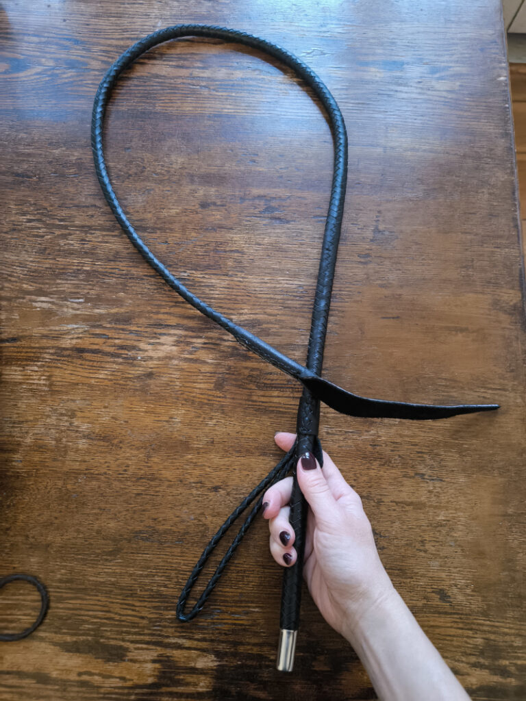 A hand holding a black braided leather whip on a wooden table.