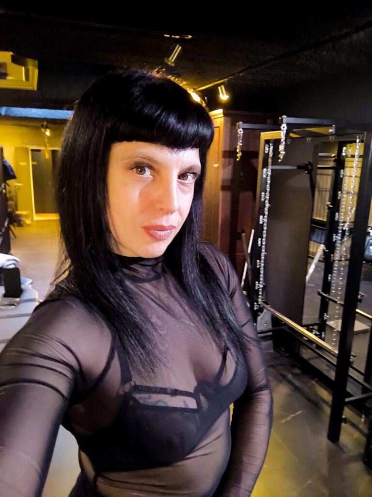 A person in a black mesh top taking a self-portrait in a dungeon studio with BDSM equipment in the background.
