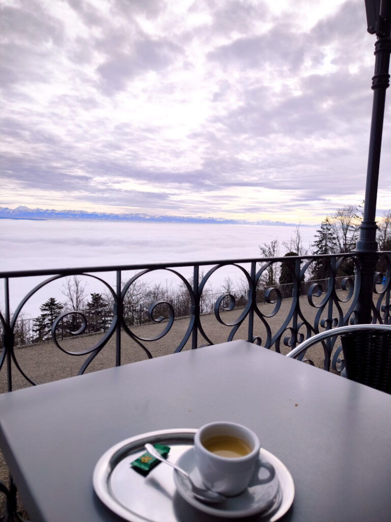 A cup of espresso on a terrace table overlooking a cloud-filled valley and distant mountains.