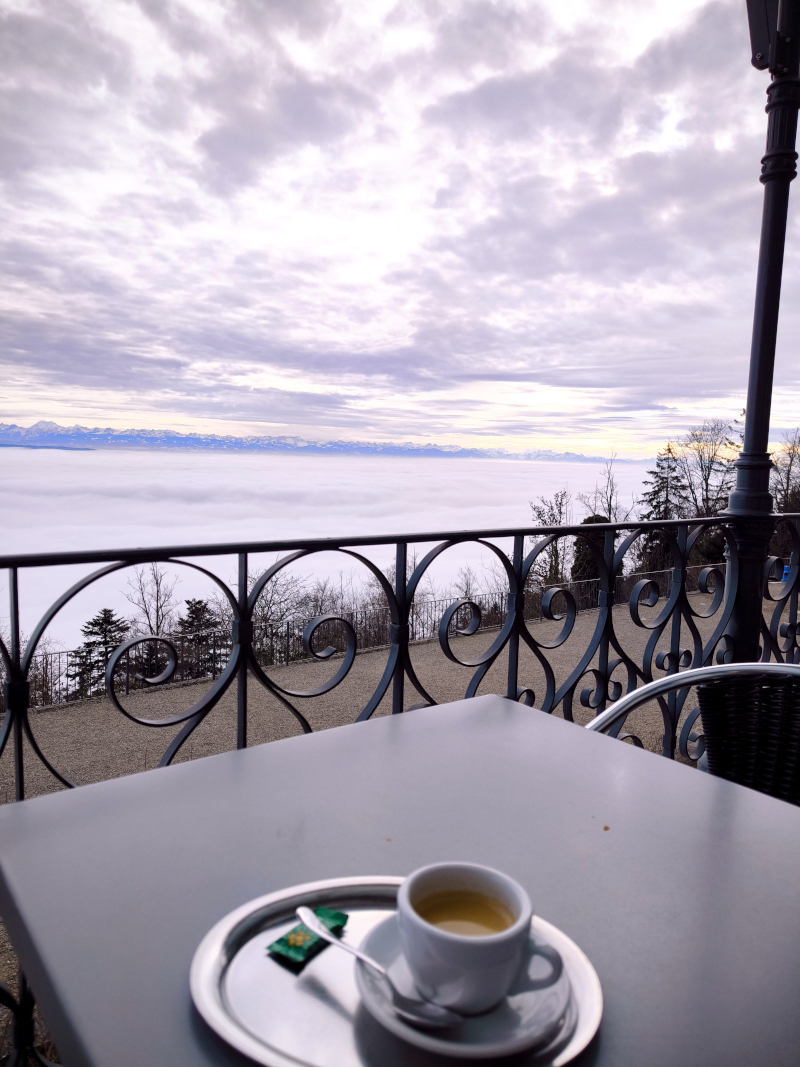 A cup of espresso on a terrace table overlooking a cloud-filled valley and distant mountains.