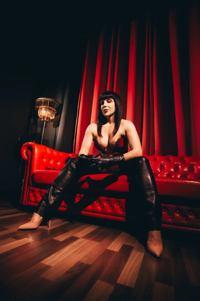 Berlin Domina Lady Vyra in a red leather corset and black leather pants, seated against red curtains